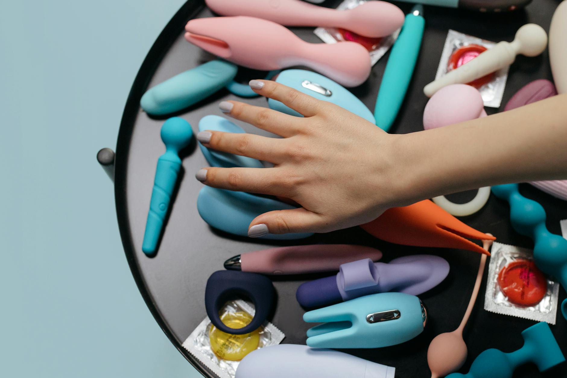 A hand reaching over a variety of colorful sex toys including lemon clitoral vibrators arranged on a table