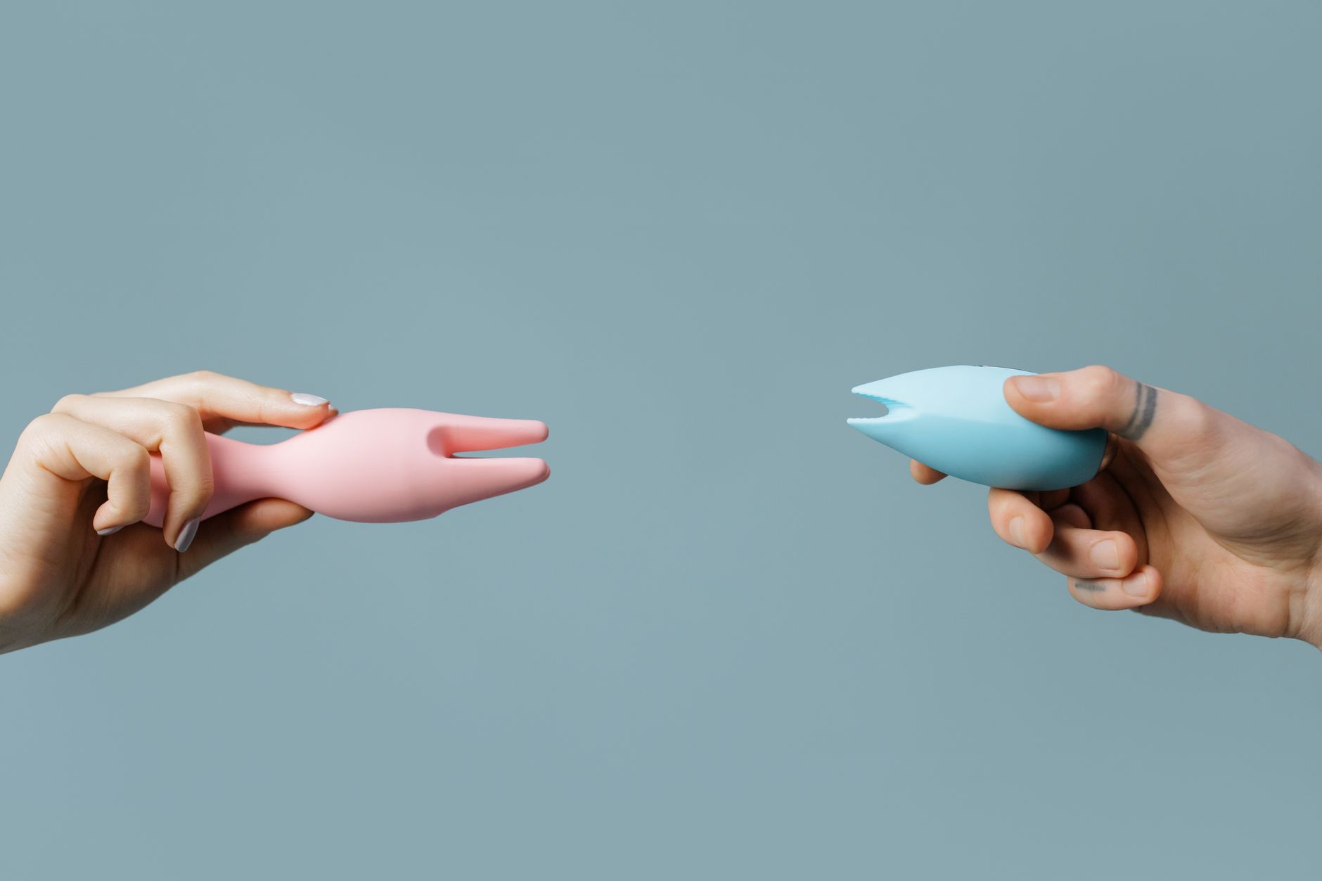 Two hands holding pink and blue silicone clitoral vibrators against a soft pastel background