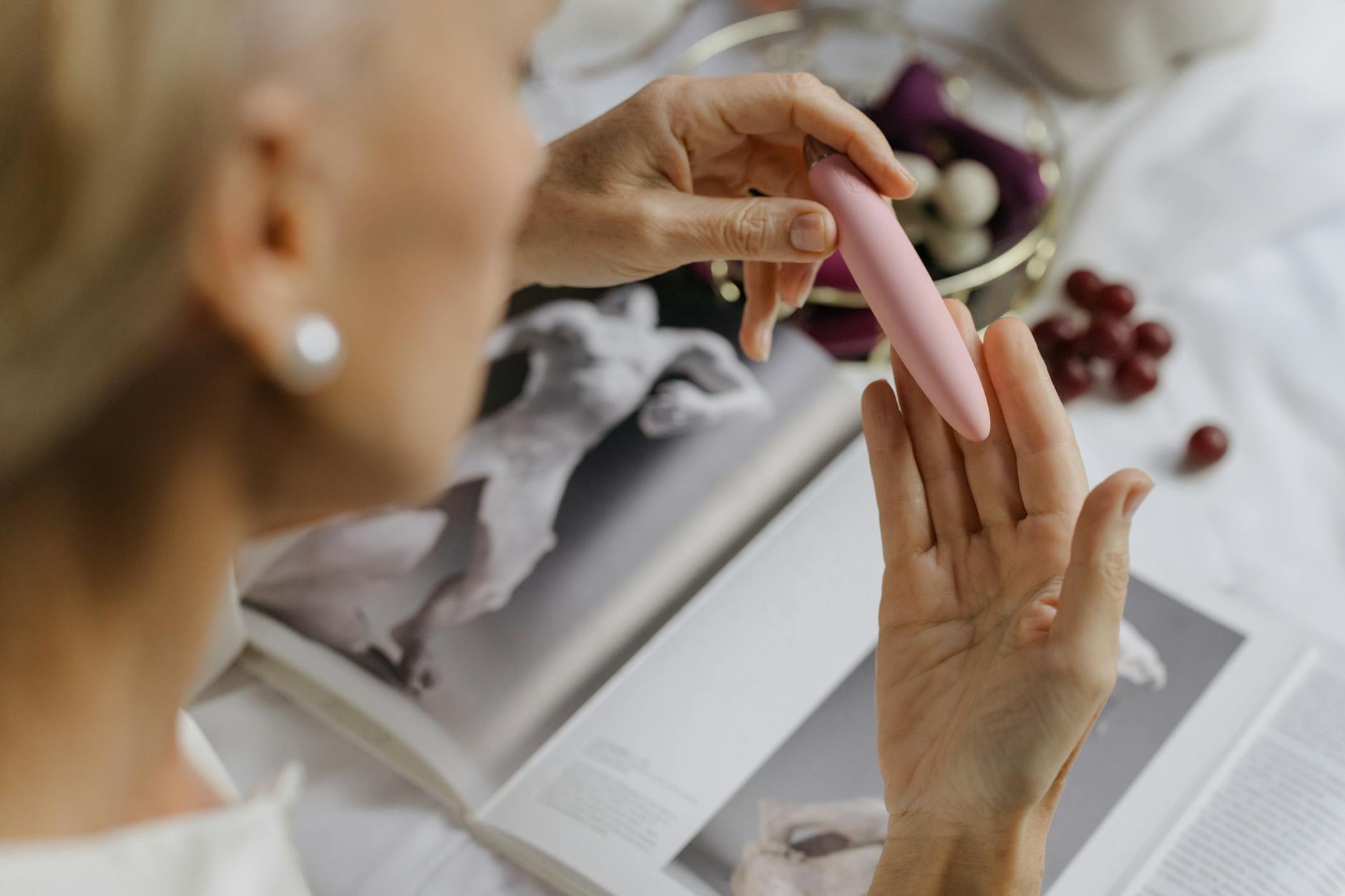 A hand holding a pink vibrator with art books and personal items in soft focus, conveying intimacy and self-discovery