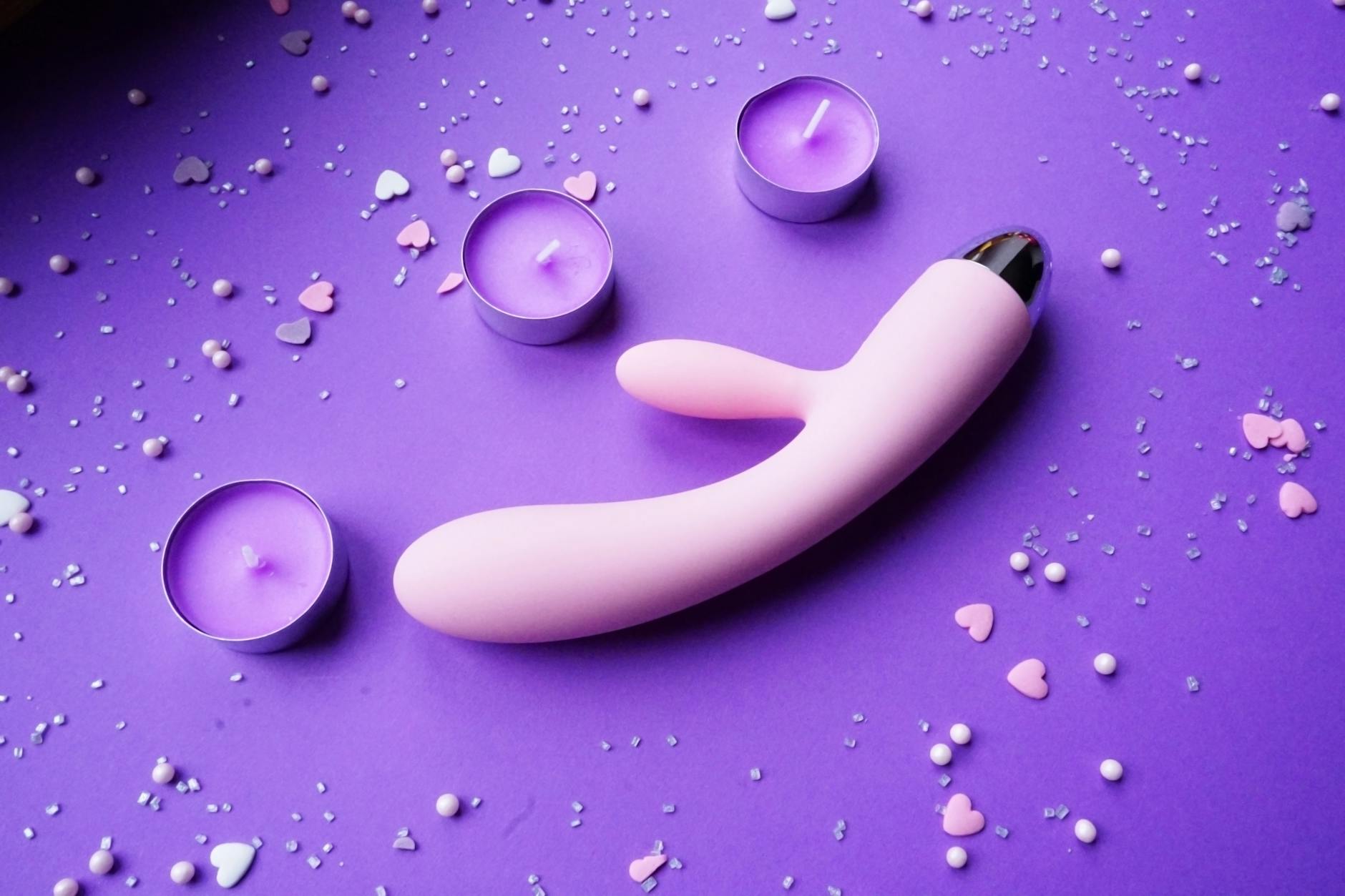 A pink vibrator surrounded by heart confetti and candles on a purple background, representing desire and sensual pleasure.